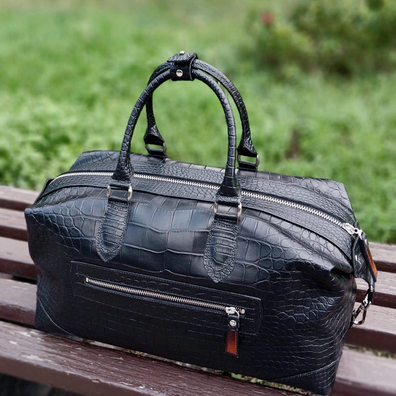May include: A black duffel bag with a textured crocodile skin pattern, featuring two top handles, a main zipper closure, and a horizontal zippered pocket on the front. The bag has silver-toned hardware and a rectangular shape.
