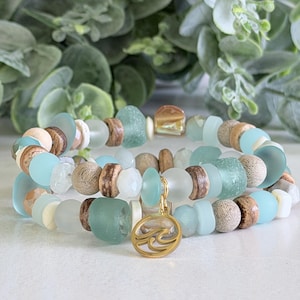 May include: A multi-strand bracelet featuring a mix of light blue and seafoam green frosted glass beads, textured brown wooden beads, and small white spacers. A circular gold-toned metal charm with a stylized wave design hangs from the center.