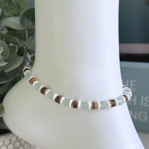 May include: An anklet featuring a repeating pattern of light seafoam green round beads, brown textured cylindrical beads, and thin white flat disc spacers. The beads are strung together in a consistent sequence to form a circular band.