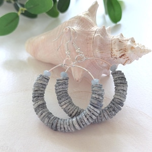 May include: A pair of hoop earrings featuring silver-toned wire hooks and circular frames. Each earring is adorned with a series of small, flat, grey, textured stone discs and a single light blue bead at the top of the hoop structure.