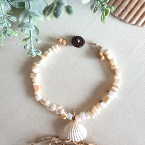 May include: A beaded bracelet featuring small, square-cut off-white shell beads strung on a cord. The bracelet includes a single white ribbed seashell charm at the center and is secured with a dark brown circular button closure and small wooden beads.