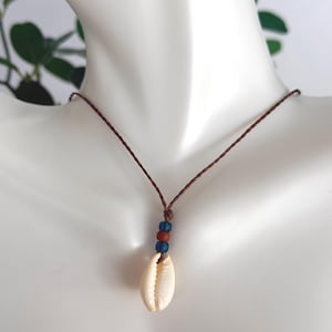 May include: A necklace featuring a single white cowrie shell pendant suspended from a thin brown braided cord. The pendant is accented with three small beads in alternating blue and terracotta colors positioned directly above the shell.