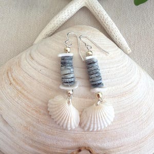 May include: A pair of dangle earrings featuring silver-toned hooks, small metallic beads, stacks of flat grey circular discs, white spacers, and a single white fan-shaped seashell pendant hanging from the base of each earring component.