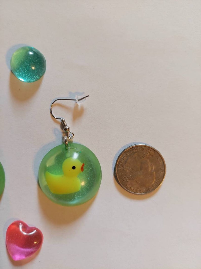 May include: A pair of green and yellow earrings with a small yellow rubber ducky inside a clear green resin circle. The earrings are hanging from silver hooks.