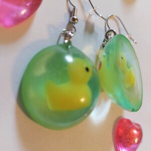 May include: A pair of green and yellow resin earrings with a small yellow rubber ducky inside each earring. The earrings are on a silver hook.