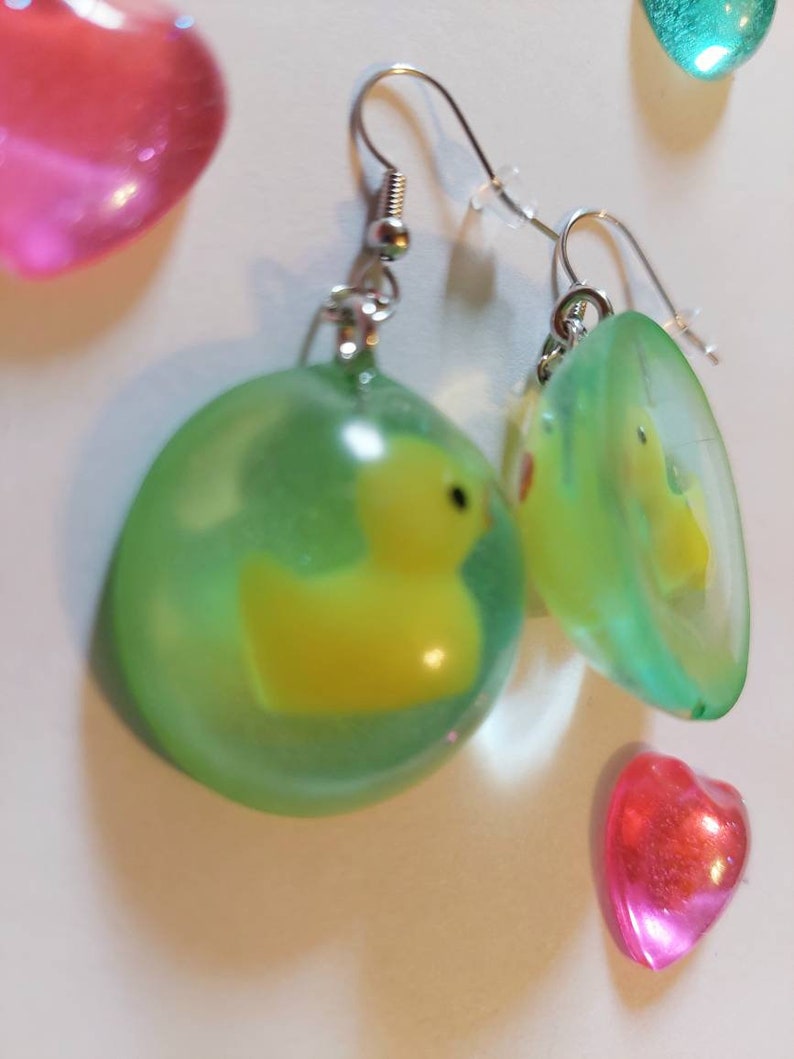 May include: A pair of green and yellow resin earrings with a small yellow rubber ducky inside each earring. The earrings are on a silver hook.