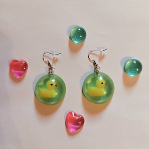 May include: A pair of green resin earrings with a yellow rubber ducky inside each earring. The earrings are on a white surface with two pink hearts and two green glass stones.