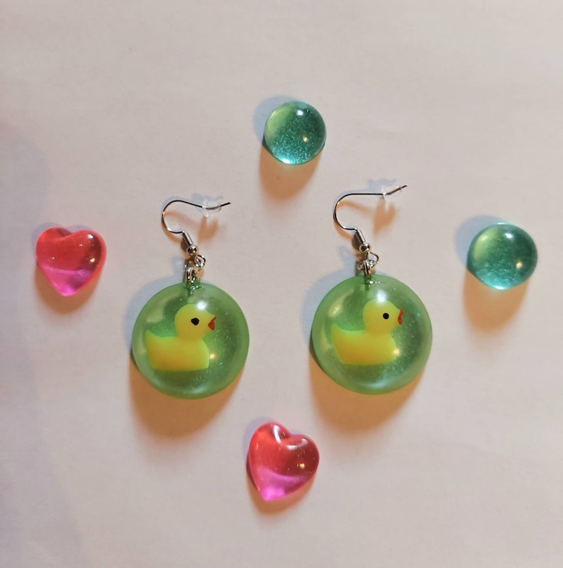 May include: A pair of green resin earrings with a yellow rubber ducky inside each earring. The earrings are on a white surface with two pink hearts and two green glass stones.