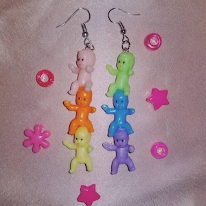 以下が含まれることがあります： カラフルなプラスチック製の赤ちゃんの人形が積み重なった、ユニークなイヤリングのペア。各イヤリングは異なる配色で、ピンク、オレンジ、黄色、紫、緑、青の赤ちゃんがいます。イヤリングはシルバーカラーのフックから吊り下げられています。