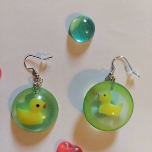 May include: A pair of green and yellow earrings with a rubber ducky inside each earring. The earrings are on a white background.