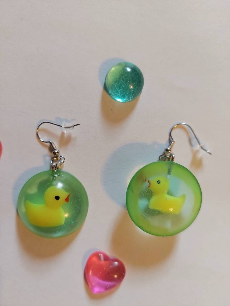 May include: A pair of green and yellow earrings with a rubber ducky inside each earring. The earrings are on a white background.