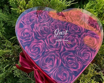 Può includere: Una scatola a forma di cuore piena di rose di seta rosa scuro. La scatola è trasparente e ha un nastro rosso legato intorno. Il testo "Just for you with love" è stampato sulla scatola.