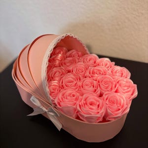 May include: A pink baby crib shaped box filled with pink satin roses. The box has a white lace trim and a white satin bow.