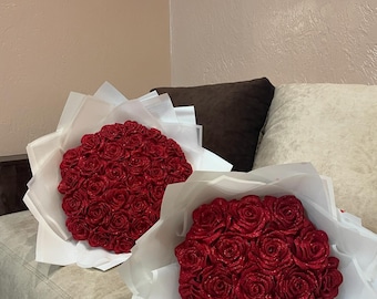 May include: Two bouquets of red glitter roses wrapped in white tissue paper. The roses are arranged in a circular shape and are a deep red color.