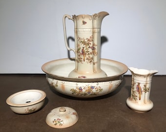 Op de afbeelding: Een vintage badkamerset met een kan, een kom en twee kleinere containers. De set is versierd met een bloemenpatroon en vogels.