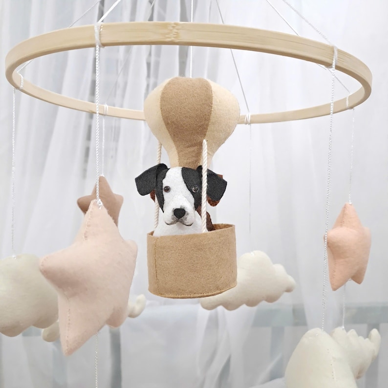 May include: A wooden baby mobile with a brown and tan hot air balloon, a white and black dog, and pink and white stars. The mobile is hanging from a white ceiling.
