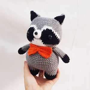 Puede incluir: Un juguete de peluche de mapache gris tejido a crochet con ojos negros, nariz blanca y orejas negras. El juguete lleva una pajarita naranja.