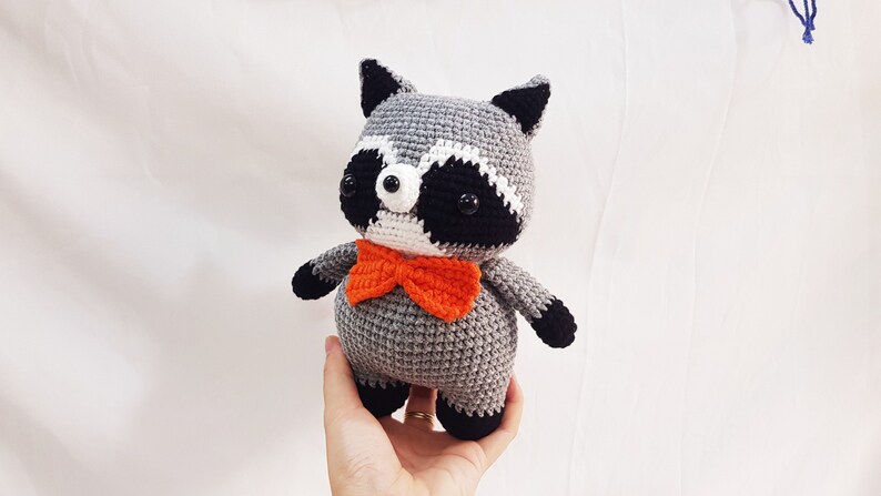 Puede incluir: Un juguete de peluche de mapache gris tejido a crochet con ojos negros, nariz blanca y orejas negras. El juguete lleva una pajarita naranja.
