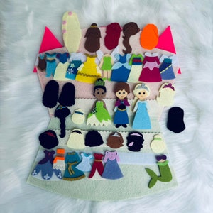 Op de afbeelding: Een vilt aankleedset met prinsessen thema outfits, kapsels en karakter figuren. De set bevat jurken in blauw, geel, roze en groen, met accessoires en karakter uitsparingen. De achtergrond is een zacht, wit, pluizig oppervlak.