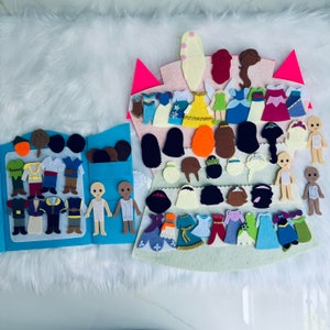 Op de afbeelding: Een vilten verkleedset met een blauwe, boekachtige basis en een kasteelvormige achtergrond. De set bevat vilten poppen, diverse outfits en kapsels in meerdere kleuren. De achtergrond is een zacht, wit oppervlak.