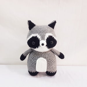 Puede incluir: Un juguete de peluche de mapache tejido a crochet con ojos negros, nariz blanca y pelaje gris y blanco.
