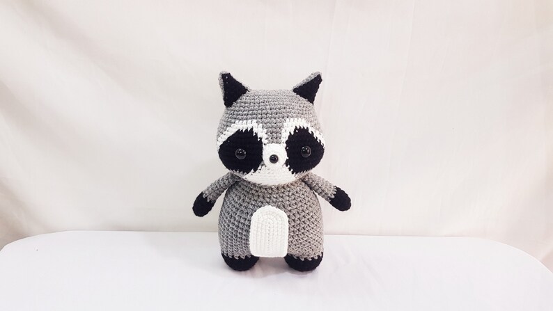 Puede incluir: Un juguete de peluche de mapache tejido a crochet con ojos negros, nariz blanca y pelaje gris y blanco.