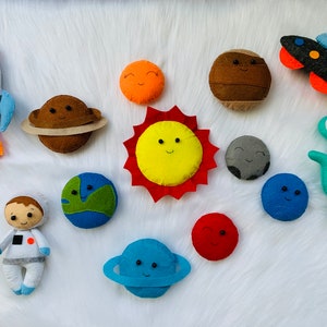 May include: A set of felt planets and space-themed characters, including a rocket, astronaut, alien, and a sun with a smiling face. The planets are in various colours, including blue, green, red, brown, and grey.