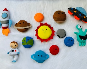 Puede incluir: Un conjunto de planetas de fieltro y personajes con temática espacial, que incluyen un cohete, un astronauta, un extraterrestre y un sol con una cara sonriente. Los planetas son de varios colores, incluyendo azul, verde, rojo, marrón y gris.