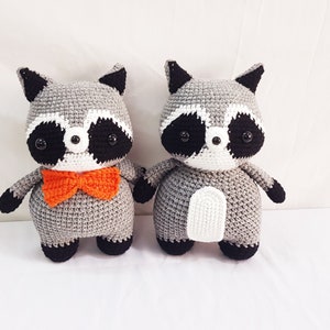 Puede incluir: Dos peluches de mapache tejidos a crochet, uno con una pajarita naranja brillante. Ambos tienen ojos negros, narices blancas y pelaje gris y blanco.