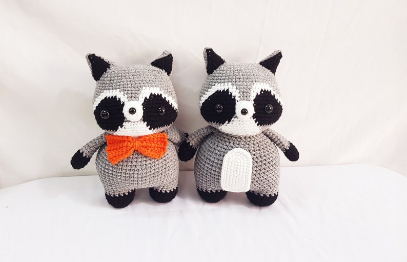 Puede incluir: Dos peluches de mapache tejidos a crochet, uno con una pajarita naranja brillante. Ambos tienen ojos negros, narices blancas y pelaje gris y blanco.