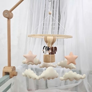 May include: A wooden baby mobile with a hot air balloon, two dogs, and clouds. The balloon is brown and yellow, the dogs are brown and white, and the clouds are white.