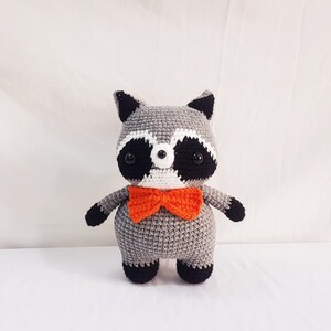 Puede incluir: Un juguete de peluche de mapache gris tejido a crochet con ojos negros, nariz blanca y orejas negras. El juguete lleva una pajarita naranja.