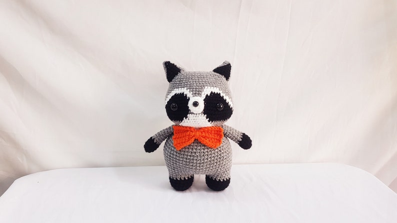 Puede incluir: Un juguete de peluche de mapache gris tejido a crochet con ojos negros, nariz blanca y orejas negras. El juguete lleva una pajarita naranja.