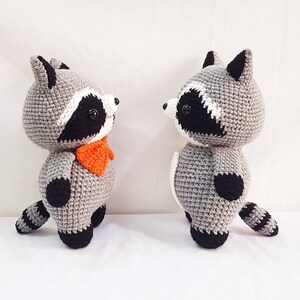 Puede incluir: Dos peluches de mapache tejidos a crochet, uno con una corbata de lazo naranja brillante, de pie uno frente al otro sobre un fondo blanco.
