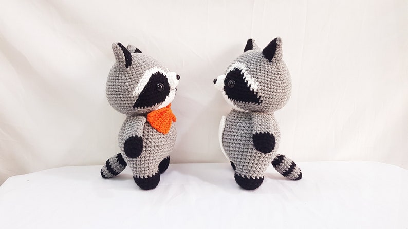 Puede incluir: Dos peluches de mapache tejidos a crochet, uno con una corbata de lazo naranja brillante, de pie uno frente al otro sobre un fondo blanco.