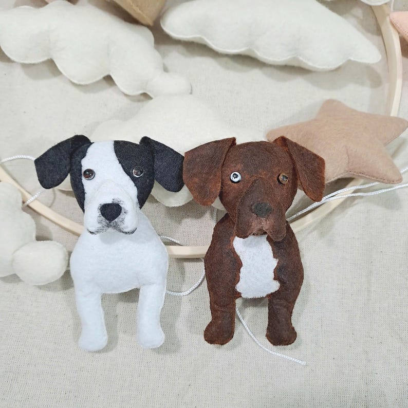 May include: Two felt dog ornaments, one black and white and one brown with white accents, hanging from a wooden ring. The ornaments are part of a mobile.