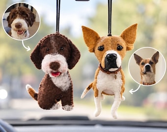 Op de afbeelding: Twee vilten hondenornamenten hangend aan een achteruitkijkspiegel. Het linker ornament is een bruin-witte hond met een rode tong. Het rechter ornament is een beige-witte hond met een bruine halsband. Elk is gekoppeld aan een ronde foto van de echte hond.
