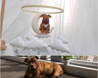 May include: A baby mobile featuring a brown dog plush toy wearing a red and black checkered scarf, seated on a crescent moon. The mobile includes white cloud shapes and a wooden ring.