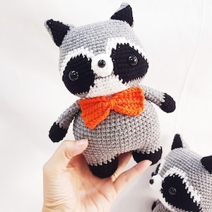 Puede incluir: Dos peluches de mapache tejidos a crochet, uno con una pajarita naranja. Los peluches son grises con detalles negros y blancos alrededor de los ojos.