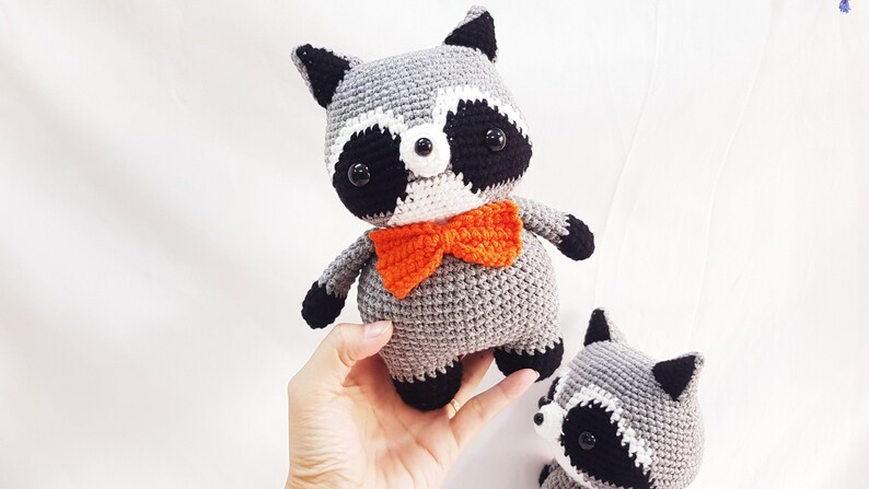 Puede incluir: Dos peluches de mapache tejidos a crochet, uno con una pajarita naranja. Los peluches son grises con detalles negros y blancos alrededor de los ojos.