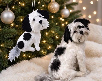 May include: A felt dog ornament featuring a white body with black spots, black ears, and a grey tail, hanging by a white string. Beside it sits a real dog with similar black and white fur, featuring a fluffy coat and a black nose and ears.