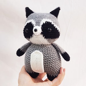 Puede incluir: Un juguete de peluche de mapache tejido a crochet con pelaje gris, blanco y negro. El juguete tiene ojos negros y una nariz negra.