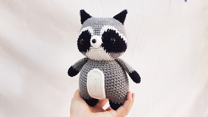 Puede incluir: Un juguete de peluche de mapache tejido a crochet con pelaje gris, blanco y negro. El juguete tiene ojos negros y una nariz negra.
