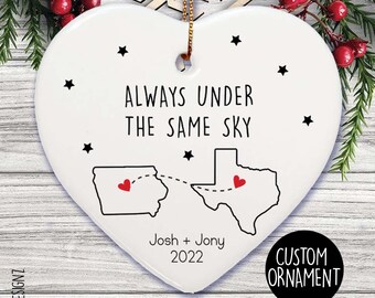 Puede incluir: Un adorno blanco en forma de corazón con el texto "ALWAYS UNDER THE SAME SKY" y dos contornos de estados conectados por una línea punteada. El adorno incluye los nombres "Josh + Jony" y el año "2022". Un círculo negro con "CUSTOM ORNAMENT" está en la parte inferior derecha.