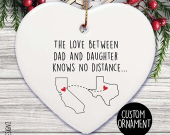 Puede incluir: Adorno blanco en forma de corazón con el texto "THE LOVE BETWEEN DAD AND DAUGHTER KNOWS NO DISTANCE..." y contornos de California y Texas con iconos de corazón rojo. Una cuerda dorada está unida para colgar.