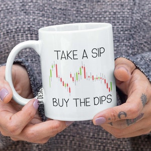 May include: White ceramic mug with a black handle. The mug has a black graphic of a stock market chart with the text "Take a Sip Buy the Dips".