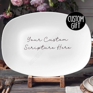 Puede incluir: Plato de servir ovalado blanco con el texto "Your Custom Scripture Here" en escritura cursiva negra. El plato está sobre un soporte de madera. El texto "CUSTOM GIFT" está en un círculo negro en la esquina superior derecha de la imagen.