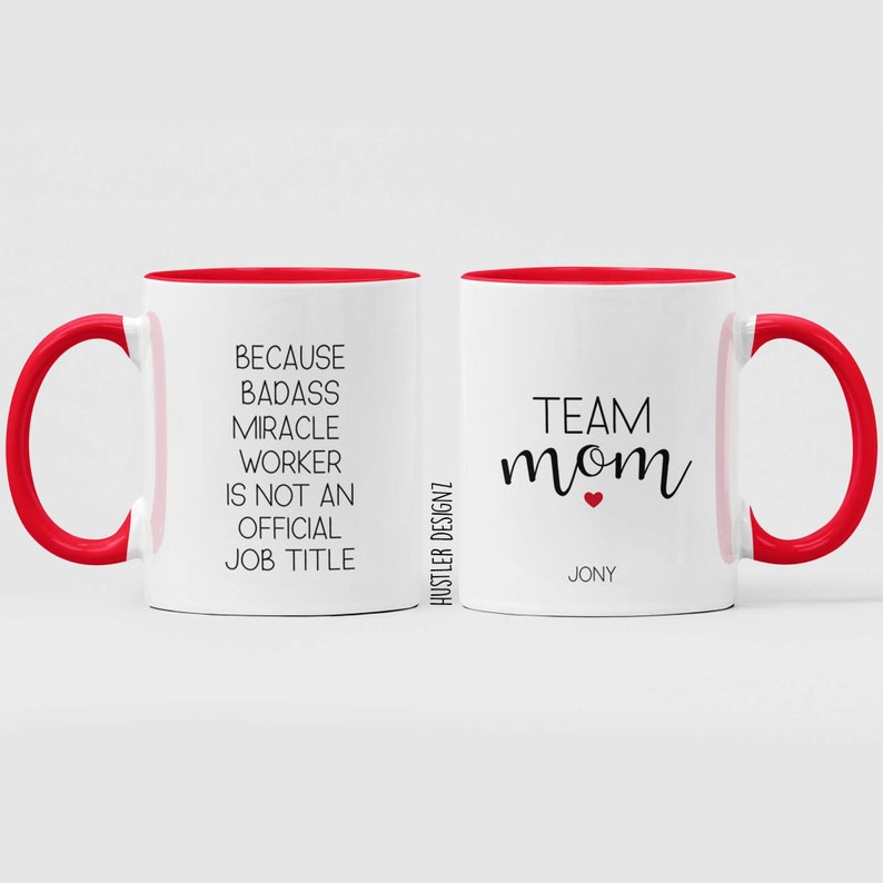 May include: White ceramic mug with a red rim and handle. The mug has black text that reads "Because Badass Miracle Worker Is Not An Official Job Title" on one side and "Team Mom" with a red heart on the other side. The text "Jony" is printed below the heart.