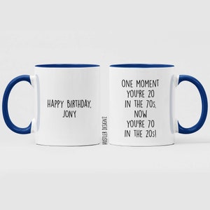 Może przedstawiać: Biały kubek ceramiczny z niebieskim rantem i uchwytem. Kubek ma napis "Happy Birthday, Jony" po jednej stronie i "One moment you're 20 in the 70s. Now you're 70 in the 20s!" po drugiej.
