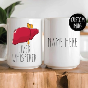 Könnte beinhalten: Zwei weiße Keramikbecher. Ein Becher zeigt eine rote Leberillustration mit dem Text "LIVER WHISPERER". Der andere Becher hat den Text "NAME HERE" und einen schwarzen Kreis mit "CUSTOM MUG".